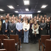 magem mostra um grupo numeroso de pessoas posando para uma foto em um audit&oacute;rio. Os participantes est&atilde;o organizados em filas, &agrave; frente de cadeiras de madeira alinhadas, com um corredor central. Ao fundo, h&aacute; uma tela de proje&ccedil;&atilde;o com um logotipo institucional do governo do Rio Grande do Sul.
No ambiente, predominam tons escuros no teto e paredes, com ilumina&ccedil;&atilde;o de pain&eacute;is retangulares. As pessoas vestem roupas formais e informais, algumas com crach&aacute;s, sugerindo um evento profissional ou institucional. H&aacute; uma mesa lateral com objetos e uma garrafa de &aacute;gua vis&iacute;vel no ch&atilde;o, pr&oacute;xima &agrave;s cadeiras.