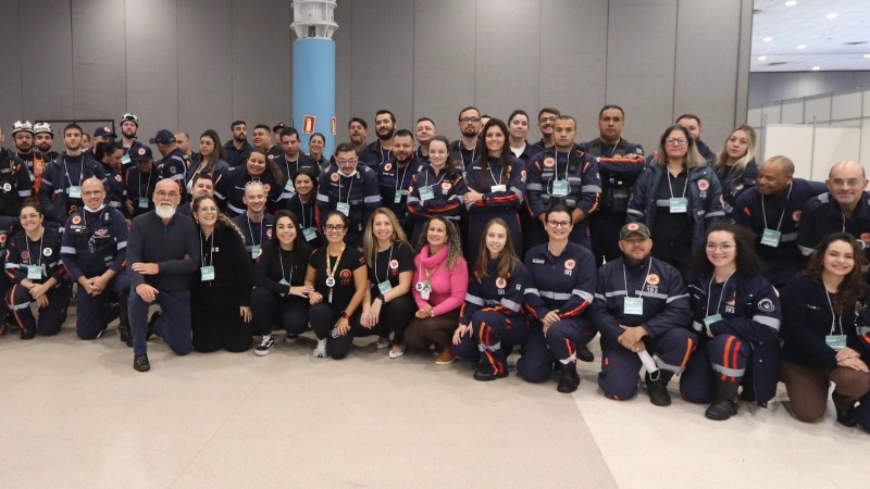 Fotografia de grupo em ambiente interno amplo, com piso claro e paredes cinza lisas. Ao fundo, h&aacute; uma grande coluna central azul-clara e ilumina&ccedil;&atilde;o artificial uniforme no teto, t&iacute;pica de centros de eventos ou pavilh&otilde;es.
No primeiro plano e no centro da imagem, cerca de v&aacute;rias dezenas de pessoas posam reunidas para uma foto coletiva. Parte do grupo est&aacute; agachada ou ajoelhada na frente, enquanto outra parte permanece em p&eacute; atr&aacute;s, formando duas fileiras. Todas as pessoas est&atilde;o voltadas em dire&ccedil;&atilde;o &agrave; c&acirc;mera.
A maioria veste uniformes escuros com detalhes refletivos e faixas em laranja, caracter&iacute;sticos de equipes de atendimento de emerg&ecirc;ncia, com logotipos do SAMU (Servi&ccedil;o de Atendimento M&oacute;vel de Urg&ecirc;ncia). Algumas pessoas usam crach&aacute;s pendurados no pesco&ccedil;o, indicando participa&ccedil;&atilde;o em evento ou competi&ccedil;&atilde;o. H&aacute; tamb&eacute;m algumas pessoas com roupas casuais entre os uniformes.
Os uniformes exibem elementos como bras&otilde;es e inscri&ccedil;&otilde;es &ldquo;192&rdquo;, al&eacute;m de patches com bandeiras e s&iacute;mbolos institucionais. Alguns integrantes possuem acess&oacute;rios como bon&eacute;s ou equipamentos presos ao uniforme.
O grupo est&aacute; organizado de maneira compacta, ocupando praticamente toda a largura da imagem. O espa&ccedil;o ao redor &eacute; amplo e sem muitos objetos vis&iacute;veis, refor&ccedil;ando o car&aacute;ter institucional ou de evento.
A cena sugere o registro de uma equipe ou de participantes de uma atividade coletiva, possivelmente relacionada a treinamento, competi&ccedil;&atilde;o ou evento na &aacute;rea de atendimento de urg&ecirc;ncia e sa&uacute;de.