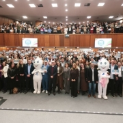 Fotografia em plano aberto de um audit&oacute;rio amplo com paredes revestidas de madeira e ilumina&ccedil;&atilde;o forte no teto. O espa&ccedil;o est&aacute; completamente cheio, com centenas de pessoas distribu&iacute;das em dois n&iacute;veis: plateia inferior e balc&atilde;o superior.
Na parte frontal, um grande grupo est&aacute; posicionado lado a lado, voltado para a c&acirc;mera, segurando certificados ou folhas de papel. Entre essas pessoas, h&aacute; dois mascotes em formato de personagens brancos, com apar&ecirc;ncia arredondada, associados ao SUS; um deles possui a sigla &ldquo;SUS&rdquo; vis&iacute;vel no corpo.
Ao fundo, nas fileiras elevadas e no mezanino, mais participantes tamb&eacute;m seguram documentos semelhantes, formando um conjunto denso e uniforme de pessoas. Muitos est&atilde;o vestidos com roupas formais ou semiformais, como blazers, camisas e cal&ccedil;as sociais.
No centro do audit&oacute;rio, acima da plateia inferior, h&aacute; dois pain&eacute;is ou telas com um logotipo institucional colorido, embora o texto n&atilde;o seja completamente leg&iacute;vel na imagem.
O piso pr&oacute;ximo &agrave; c&acirc;mera &eacute; cinza, com uma faixa iluminada delimitando a &aacute;rea frontal. A composi&ccedil;&atilde;o geral transmite um evento coletivo de reconhecimento ou certifica&ccedil;&atilde;o, com grande participa&ccedil;&atilde;o e organiza&ccedil;&atilde;o em grupo.