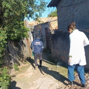 Duas pessoas caminham por um beco estreito entre casas, com muros de tijolo e vegeta&ccedil;&atilde;o ao redor. Uma veste jaleco e a outra casaco com identifica&ccedil;&atilde;o, sugerindo visita ou atendimento em &aacute;rea residencial.
