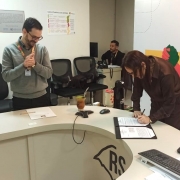 Uma pessoa assina um documento sobre uma mesa institucional enquanto outra observa ao lado; h&aacute; cadeiras, computador e logotipo do RS ao fundo.