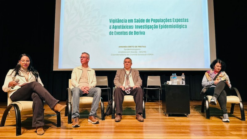 A imagem mostra um palco de audit&oacute;rio durante um semin&aacute;rio, com quatro participantes sentados em poltronas. Ao fundo, h&aacute; um tel&atilde;o com o t&iacute;tulo &ldquo;III Semin&aacute;rio VSPEA &ndash; 09 e 10 de junho &ndash; Porto Alegre/RS&rdquo; e o tema da apresenta&ccedil;&atilde;o, que aborda vigil&acirc;ncia em sa&uacute;de de popula&ccedil;&otilde;es expostas a agrot&oacute;xicos. Tamb&eacute;m aparece o nome da palestrante e sua vincula&ccedil;&atilde;o institucional. No centro do palco h&aacute; uma pequena mesa com garrafas de &aacute;gua, e o ambiente possui ilumina&ccedil;&atilde;o focada nos participantes, com piso de madeira e fundo escuro.