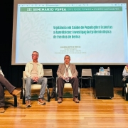 A imagem mostra um palco de audit&oacute;rio durante um semin&aacute;rio, com quatro participantes sentados em poltronas. Ao fundo, h&aacute; um tel&atilde;o com o t&iacute;tulo &ldquo;III Semin&aacute;rio VSPEA &ndash; 09 e 10 de junho &ndash; Porto Alegre/RS&rdquo; e o tema da apresenta&ccedil;&atilde;o, que aborda vigil&acirc;ncia em sa&uacute;de de popula&ccedil;&otilde;es expostas a agrot&oacute;xicos. Tamb&eacute;m aparece o nome da palestrante e sua vincula&ccedil;&atilde;o institucional. No centro do palco h&aacute; uma pequena mesa com garrafas de &aacute;gua, e o ambiente possui ilumina&ccedil;&atilde;o focada nos participantes, com piso de madeira e fundo escuro.