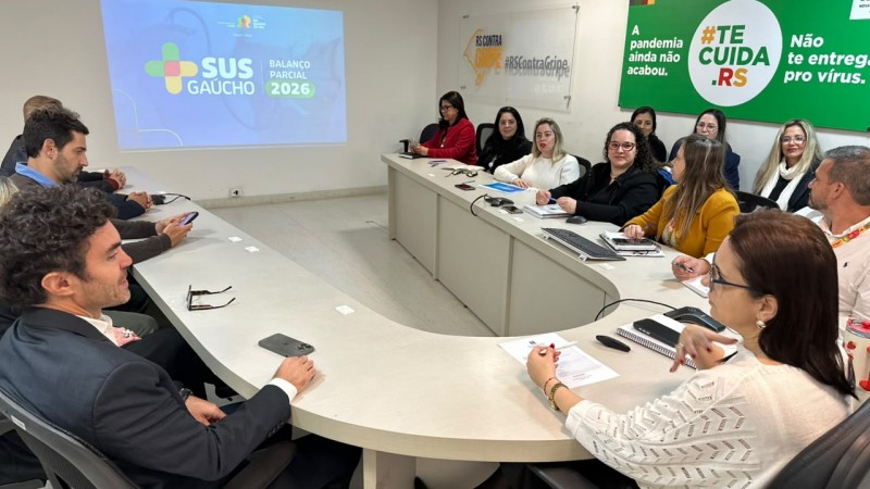 Reuni&atilde;o institucional em sala de confer&ecirc;ncias, com cerca de dez pessoas sentadas ao redor de uma mesa, acompanhando uma apresenta&ccedil;&atilde;o projetada na parede com o t&iacute;tulo &ldquo;SUS Ga&uacute;cho &ndash; Balan&ccedil;o Parcial 2026&rdquo;. No ambiente, h&aacute; computadores, celulares e anota&ccedil;&otilde;es sobre a mesa, al&eacute;m de cartazes institucionais nas paredes relacionados &agrave; sa&uacute;de p&uacute;blica e campanhas estaduais.