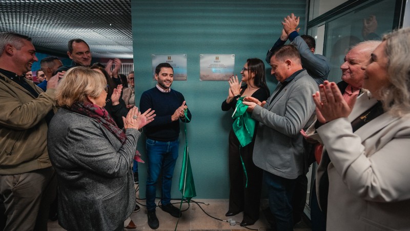 Imagem de um grupo de v&aacute;rias pessoas reunidas em um ambiente interno, em torno de duas placas comemorativas fixadas em uma parede. As placas possuem texto (n&atilde;o totalmente leg&iacute;vel na imagem) e parecem marcar uma inaugura&ccedil;&atilde;o. Algumas pessoas est&atilde;o pr&oacute;ximas das placas segurando um tecido verde, parcialmente removido, sugerindo o momento de descerramento. Outras pessoas ao redor est&atilde;o com as m&atilde;os erguidas, em atitude de aplauso. O espa&ccedil;o apresenta ilumina&ccedil;&atilde;o artificial e paredes em tonalidade esverdeada. As pessoas vestem roupas variadas, incluindo casacos e pe&ccedil;as formais. No centro da cena, destaca-se a &aacute;rea das placas e o tecido verde retirado, indicando um evento oficial de entrega ou inaugura&ccedil;&atilde;o de instala&ccedil;&otilde;es.