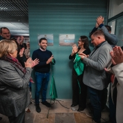 Imagem de um grupo de v&aacute;rias pessoas reunidas em um ambiente interno, em torno de duas placas comemorativas fixadas em uma parede. As placas possuem texto (n&atilde;o totalmente leg&iacute;vel na imagem) e parecem marcar uma inaugura&ccedil;&atilde;o. Algumas pessoas est&atilde;o pr&oacute;ximas das placas segurando um tecido verde, parcialmente removido, sugerindo o momento de descerramento. Outras pessoas ao redor est&atilde;o com as m&atilde;os erguidas, em atitude de aplauso. O espa&ccedil;o apresenta ilumina&ccedil;&atilde;o artificial e paredes em tonalidade esverdeada. As pessoas vestem roupas variadas, incluindo casacos e pe&ccedil;as formais. No centro da cena, destaca-se a &aacute;rea das placas e o tecido verde retirado, indicando um evento oficial de entrega ou inaugura&ccedil;&atilde;o de instala&ccedil;&otilde;es.