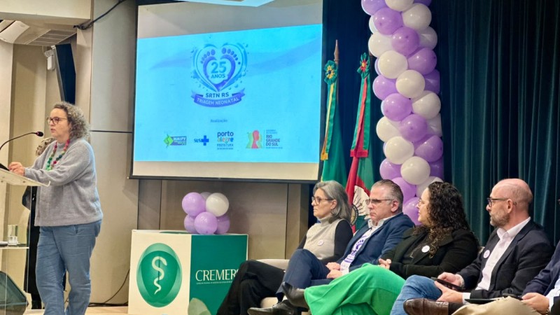 Evento realizado em audit&oacute;rio, com uma pessoa discursando em p&uacute;lpito com microfone. Ao fundo, um tel&atilde;o exibe &ldquo;25 anos SRTN RS Triagem Neonatal&rdquo; com logotipos institucionais. &Agrave; direita, um grupo de pessoas est&aacute; sentado em cadeiras, e h&aacute; decora&ccedil;&atilde;o com bal&otilde;es nas cores branco e lil&aacute;s, al&eacute;m de bandeiras.