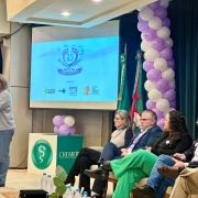 Evento realizado em audit&oacute;rio, com uma pessoa discursando em p&uacute;lpito com microfone. Ao fundo, um tel&atilde;o exibe &ldquo;25 anos SRTN RS Triagem Neonatal&rdquo; com logotipos institucionais. &Agrave; direita, um grupo de pessoas est&aacute; sentado em cadeiras, e h&aacute; decora&ccedil;&atilde;o com bal&otilde;es nas cores branco e lil&aacute;s, al&eacute;m de bandeiras.