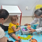Ambiente interno semelhante a sala de atendimento infantil. No ch&atilde;o, h&aacute; um tapete colorido com ilustra&ccedil;&otilde;es, onde uma crian&ccedil;a pequena est&aacute; sentada, cercada por brinquedos diversos, como pe&ccedil;as de encaixe, animais de pl&aacute;stico e um objeto em formato de trilho ou caminho colorido.
&Agrave; frente da crian&ccedil;a, uma pessoa adulta vestindo jaleco branco est&aacute; ajoelhada e segura uma caixa pl&aacute;stica contendo v&aacute;rios brinquedos, colocando-os ao alcance da crian&ccedil;a. Ao lado esquerdo, outra pessoa adulta sentada observa a intera&ccedil;&atilde;o.
Ao fundo, h&aacute; m&oacute;veis simples, como uma mesa pequena com bordas coloridas e cadeiras infantis, al&eacute;m de uma tomada el&eacute;trica na parede. O ambiente &eacute; organizado e voltado ao atendimento infantil, sugerindo um contexto de avalia&ccedil;&atilde;o, cuidado ou acompanhamento de desenvolvimento.