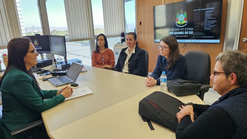 Imagem de uma reuni&atilde;o realizada em uma sala administrativa ampla e iluminada por grandes janelas com persianas verticais. Cinco pessoas est&atilde;o sentadas ao redor de uma mesa retangular clara. Sobre a mesa h&aacute; um notebook aberto, monitores de computador, garrafas de &aacute;gua, documentos e bolsas.

Na parede ao fundo, uma tela exibe a identidade visual do Governo do Estado do Rio Grande do Sul e da Secretaria da Sa&uacute;de. Ao lado da tela h&aacute; revestimento em madeira e uma porta. Pela janela &eacute; poss&iacute;vel ver parte da paisagem externa e edifica&ccedil;&otilde;es ao longe.

Os participantes est&atilde;o distribu&iacute;dos em ambos os lados da mesa, voltados para a discuss&atilde;o. O ambiente possui caracter&iacute;sticas de escrit&oacute;rio institucional, com equipamentos de inform&aacute;tica, esta&ccedil;&otilde;es de trabalho e mobili&aacute;rio corporativo.
