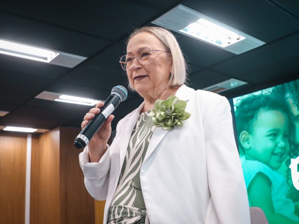 Pessoa segura um microfone durante apresentação em auditório. Veste blazer claro sobre roupa com estampa em tons de verde e usa óculos. Ao fundo, há um telão com imagem de uma criança em tons esverdeados e iluminação interna.