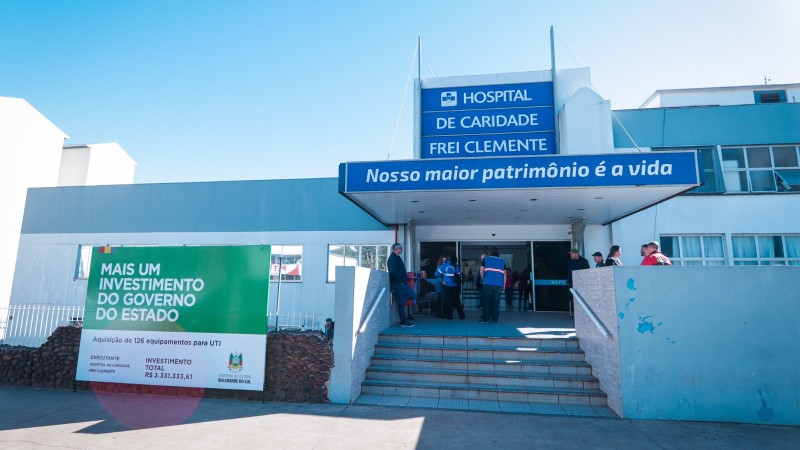 Fotografia da fachada principal do Hospital de Caridade Frei Clemente, em Soledade. O pr&eacute;dio, de cor clara, possui estrutura t&eacute;rrea com entrada central coberta por uma marquise azul. Sobre a entrada h&aacute; uma placa com os dizeres &ldquo;Hospital de Caridade Frei Clemente&rdquo; e, logo abaixo, a frase &ldquo;Nosso maior patrim&ocirc;nio &eacute; a vida&rdquo;. Em frente ao acesso principal h&aacute; uma escadaria com corrim&atilde;os laterais e v&aacute;rias pessoas distribu&iacute;das pr&oacute;ximas &agrave; entrada.

&Agrave; esquerda da imagem, destaca-se um grande painel verde e branco informando um investimento do Governo do Estado para a aquisi&ccedil;&atilde;o de 126 equipamentos para UTI. O painel menciona investimento total de R$ 2.331.333,61 e exibe as logomarcas do Hospital de Caridade Frei Clemente e do Governo do Estado do Rio Grande do Sul.

O c&eacute;u aparece limpo e azul, ocupando grande parte da parte superior da fotografia. A ilumina&ccedil;&atilde;o &eacute; diurna, com forte incid&ecirc;ncia de luz solar sobre a fachada e sobre a &aacute;rea pavimentada em frente ao hospital. A imagem apresenta uma vis&atilde;o ampla da frente da institui&ccedil;&atilde;o, evidenciando a entrada principal e o painel de investimento p&uacute;blico.