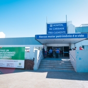 Fotografia da fachada principal do Hospital de Caridade Frei Clemente, em Soledade. O pr&eacute;dio, de cor clara, possui estrutura t&eacute;rrea com entrada central coberta por uma marquise azul. Sobre a entrada h&aacute; uma placa com os dizeres &ldquo;Hospital de Caridade Frei Clemente&rdquo; e, logo abaixo, a frase &ldquo;Nosso maior patrim&ocirc;nio &eacute; a vida&rdquo;. Em frente ao acesso principal h&aacute; uma escadaria com corrim&atilde;os laterais e v&aacute;rias pessoas distribu&iacute;das pr&oacute;ximas &agrave; entrada.

&Agrave; esquerda da imagem, destaca-se um grande painel verde e branco informando um investimento do Governo do Estado para a aquisi&ccedil;&atilde;o de 126 equipamentos para UTI. O painel menciona investimento total de R$ 2.331.333,61 e exibe as logomarcas do Hospital de Caridade Frei Clemente e do Governo do Estado do Rio Grande do Sul.

O c&eacute;u aparece limpo e azul, ocupando grande parte da parte superior da fotografia. A ilumina&ccedil;&atilde;o &eacute; diurna, com forte incid&ecirc;ncia de luz solar sobre a fachada e sobre a &aacute;rea pavimentada em frente ao hospital. A imagem apresenta uma vis&atilde;o ampla da frente da institui&ccedil;&atilde;o, evidenciando a entrada principal e o painel de investimento p&uacute;blico.