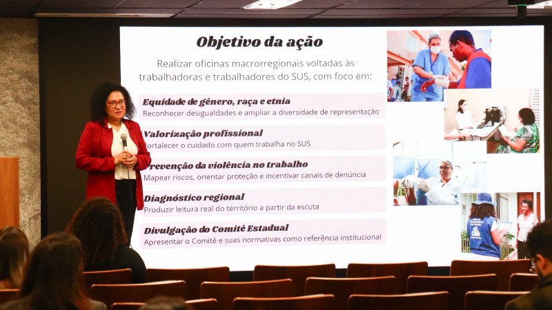 Segunda imagem
Ambiente: Audit&oacute;rio com p&uacute;blico sentado, assistindo a uma pessoa que fala diante de uma tela de proje&ccedil;&atilde;o.

Elementos: O slide projetado tem o t&iacute;tulo &ldquo;Objetivo da a&ccedil;&atilde;o&rdquo; e cont&eacute;m fotos de profissionais de sa&uacute;de em diferentes contextos.

Conte&uacute;do da tela:

Lista de objetivos das oficinas macrorregionais voltadas a trabalhadores do SUS.

T&oacute;picos incluem:

Equidade de g&ecirc;nero, ra&ccedil;a e etnia (reconhecimento das desigualdades e amplia&ccedil;&atilde;o da diversidade).

Valoriza&ccedil;&atilde;o profissional (fortalecer o cuidado com quem trabalha no SUS).

Preven&ccedil;&atilde;o da viol&ecirc;ncia no trabalho (mapear riscos, orientar prote&ccedil;&atilde;o e incentivar den&uacute;ncias).

Diagn&oacute;stico regional (leitura real do territ&oacute;rio a partir da escuta).

Divulga&ccedil;&atilde;o do Comit&ecirc; Estadual (apresenta&ccedil;&atilde;o de normativas como refer&ecirc;ncia institucional).