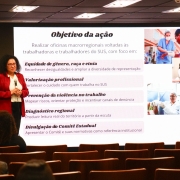 Segunda imagem
Ambiente: Audit&oacute;rio com p&uacute;blico sentado, assistindo a uma pessoa que fala diante de uma tela de proje&ccedil;&atilde;o.

Elementos: O slide projetado tem o t&iacute;tulo &ldquo;Objetivo da a&ccedil;&atilde;o&rdquo; e cont&eacute;m fotos de profissionais de sa&uacute;de em diferentes contextos.

Conte&uacute;do da tela:

Lista de objetivos das oficinas macrorregionais voltadas a trabalhadores do SUS.

T&oacute;picos incluem:

Equidade de g&ecirc;nero, ra&ccedil;a e etnia (reconhecimento das desigualdades e amplia&ccedil;&atilde;o da diversidade).

Valoriza&ccedil;&atilde;o profissional (fortalecer o cuidado com quem trabalha no SUS).

Preven&ccedil;&atilde;o da viol&ecirc;ncia no trabalho (mapear riscos, orientar prote&ccedil;&atilde;o e incentivar den&uacute;ncias).

Diagn&oacute;stico regional (leitura real do territ&oacute;rio a partir da escuta).

Divulga&ccedil;&atilde;o do Comit&ecirc; Estadual (apresenta&ccedil;&atilde;o de normativas como refer&ecirc;ncia institucional).