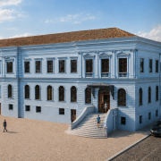Renderiza&ccedil;&atilde;o arquitet&ocirc;nica da fachada principal de um grande edif&iacute;cio hist&oacute;rico de dois pavimentos. A constru&ccedil;&atilde;o apresenta paredes azul-claras, telhado de telhas cer&acirc;micas avermelhadas, janelas altas com esquadrias escuras e elementos ornamentais cl&aacute;ssicos. No centro da fachada h&aacute; uma entrada principal elevada, acessada por uma ampla escadaria curva e coberta por uma marquise met&aacute;lica decorativa. O pr&eacute;dio ocupa uma extensa &aacute;rea urbana, cercado por &aacute;rvores, cal&ccedil;adas e vias pavimentadas. Algumas pessoas caminham em frente ao edif&iacute;cio e ve&iacute;culos est&atilde;o estacionados ao longo da rua lateral. A imagem evidencia a impon&ecirc;ncia e a simetria da arquitetura hist&oacute;rica restaurada.