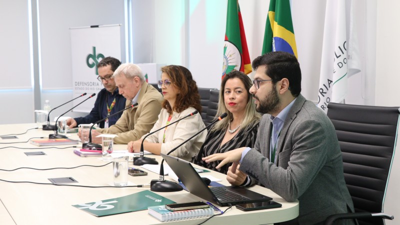 Fotografia realizada em uma sala de reuni&otilde;es institucional. Seis participantes est&atilde;o sentados lado a lado em uma mesa longa e clara, equipada com microfones, copos de &aacute;gua, cadernos, telefones celulares e um computador port&aacute;til. Em primeiro plano, uma pessoa usa o notebook enquanto faz uma exposi&ccedil;&atilde;o aos demais participantes. Ao fundo, aparecem bandeiras do Rio Grande do Sul e do Brasil, al&eacute;m de um banner com a identidade visual da Defensoria P&uacute;blica do Estado do Rio Grande do Sul. O ambiente &eacute; iluminado por luz natural proveniente de grandes janelas cobertas por persianas claras. A imagem registra um momento de apresenta&ccedil;&atilde;o t&eacute;cnica e troca de informa&ccedil;&otilde;es em contexto institucional.