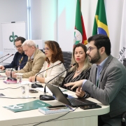 Fotografia realizada em uma sala de reuni&otilde;es institucional. Seis participantes est&atilde;o sentados lado a lado em uma mesa longa e clara, equipada com microfones, copos de &aacute;gua, cadernos, telefones celulares e um computador port&aacute;til. Em primeiro plano, uma pessoa usa o notebook enquanto faz uma exposi&ccedil;&atilde;o aos demais participantes. Ao fundo, aparecem bandeiras do Rio Grande do Sul e do Brasil, al&eacute;m de um banner com a identidade visual da Defensoria P&uacute;blica do Estado do Rio Grande do Sul. O ambiente &eacute; iluminado por luz natural proveniente de grandes janelas cobertas por persianas claras. A imagem registra um momento de apresenta&ccedil;&atilde;o t&eacute;cnica e troca de informa&ccedil;&otilde;es em contexto institucional.