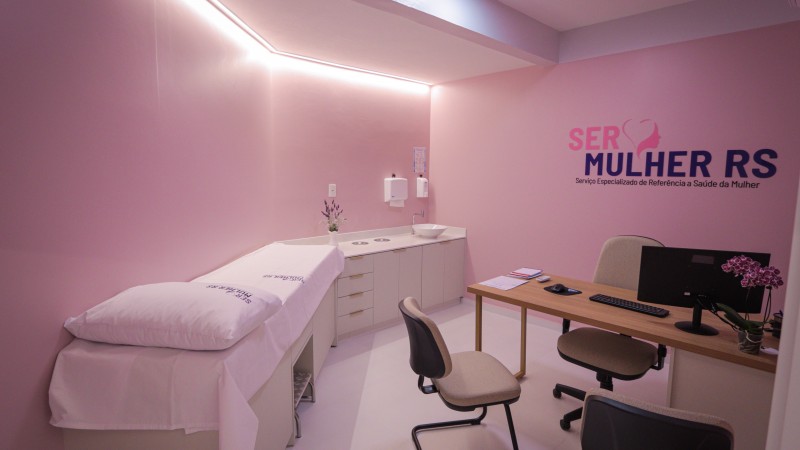 Fotografia do interior de um consult&oacute;rio do programa SERMulher RS &ndash; Servi&ccedil;o Especializado de Refer&ecirc;ncia &agrave; Sa&uacute;de da Mulher. O ambiente &eacute; compacto, organizado e predominam tons de rosa-claro nas paredes, criando uma identidade visual voltada ao atendimento em sa&uacute;de da mulher.

&Agrave; esquerda da imagem, h&aacute; uma maca de exames branca com len&ccedil;ol e travesseiro, posicionada junto &agrave; parede. Ao fundo, uma bancada branca com arm&aacute;rios inferiores abriga uma pia de apoio. Sobre a parede acima da bancada est&atilde;o instalados dispensadores de papel-toalha e sabonete, al&eacute;m de um pequeno arranjo de flores decorativas.

&Agrave; direita, encontra-se uma mesa de atendimento com acabamento em madeira clara, equipada com computador, teclado, mouse, telefone celular, documentos e um vaso com flores ornamentais. Em frente &agrave; mesa h&aacute; cadeiras destinadas ao acolhimento das pacientes.

Na parede do fundo est&aacute; aplicado o logotipo do programa, com os dizeres &ldquo;SER MULHER RS&rdquo; e a identifica&ccedil;&atilde;o &ldquo;Servi&ccedil;o Especializado de Refer&ecirc;ncia &agrave; Sa&uacute;de da Mulher&rdquo;. O espa&ccedil;o &eacute; iluminado por lumin&aacute;rias embutidas no teto e por uma faixa de ilumina&ccedil;&atilde;o indireta instalada junto ao encontro das paredes com o forro, conferindo uniformidade e conforto visual ao ambiente. A imagem evidencia um consult&oacute;rio destinado a consultas e atendimentos especializados em sa&uacute;de da mulher.