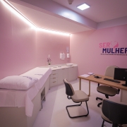 Fotografia do interior de um consult&oacute;rio do programa SERMulher RS &ndash; Servi&ccedil;o Especializado de Refer&ecirc;ncia &agrave; Sa&uacute;de da Mulher. O ambiente &eacute; compacto, organizado e predominam tons de rosa-claro nas paredes, criando uma identidade visual voltada ao atendimento em sa&uacute;de da mulher.

&Agrave; esquerda da imagem, h&aacute; uma maca de exames branca com len&ccedil;ol e travesseiro, posicionada junto &agrave; parede. Ao fundo, uma bancada branca com arm&aacute;rios inferiores abriga uma pia de apoio. Sobre a parede acima da bancada est&atilde;o instalados dispensadores de papel-toalha e sabonete, al&eacute;m de um pequeno arranjo de flores decorativas.

&Agrave; direita, encontra-se uma mesa de atendimento com acabamento em madeira clara, equipada com computador, teclado, mouse, telefone celular, documentos e um vaso com flores ornamentais. Em frente &agrave; mesa h&aacute; cadeiras destinadas ao acolhimento das pacientes.

Na parede do fundo est&aacute; aplicado o logotipo do programa, com os dizeres &ldquo;SER MULHER RS&rdquo; e a identifica&ccedil;&atilde;o &ldquo;Servi&ccedil;o Especializado de Refer&ecirc;ncia &agrave; Sa&uacute;de da Mulher&rdquo;. O espa&ccedil;o &eacute; iluminado por lumin&aacute;rias embutidas no teto e por uma faixa de ilumina&ccedil;&atilde;o indireta instalada junto ao encontro das paredes com o forro, conferindo uniformidade e conforto visual ao ambiente. A imagem evidencia um consult&oacute;rio destinado a consultas e atendimentos especializados em sa&uacute;de da mulher.