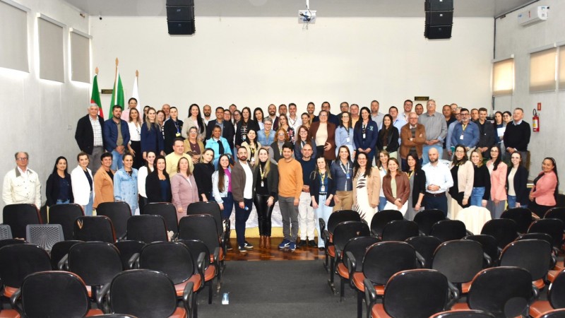 Fotografia de grupo realizada em um audit&oacute;rio. Cerca de 70 pessoas est&atilde;o posicionadas em tr&ecirc;s fileiras sobre um palco e &agrave; frente dele, voltadas para a c&acirc;mera. O grupo ocupa a parte central da imagem e re&uacute;ne participantes com diferentes trajes profissionais e casuais, muitos usando crach&aacute;s.

Em primeiro plano, h&aacute; diversas fileiras de cadeiras vazias de audit&oacute;rio, predominantemente pretas com assentos em tom alaranjado, formando um corredor central que conduz visualmente at&eacute; o grupo fotografado. O piso pr&oacute;ximo ao palco &eacute; de madeira escura.

Ao fundo, a parede &eacute; clara e sem elementos decorativos de destaque. &Agrave; esquerda do palco, aparecem bandeiras hasteadas e um p&uacute;lpito. No teto, h&aacute; caixas de som suspensas e um projetor instalado no centro. Nas laterais do audit&oacute;rio, h&aacute; janelas com persianas fechadas, al&eacute;m de aparelhos de ar-condicionado fixados nas paredes. A ilumina&ccedil;&atilde;o &eacute; uniforme e interna, t&iacute;pica de ambientes para eventos e reuni&otilde;es.