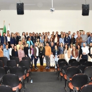 Fotografia de grupo realizada em um audit&oacute;rio. Cerca de 70 pessoas est&atilde;o posicionadas em tr&ecirc;s fileiras sobre um palco e &agrave; frente dele, voltadas para a c&acirc;mera. O grupo ocupa a parte central da imagem e re&uacute;ne participantes com diferentes trajes profissionais e casuais, muitos usando crach&aacute;s.

Em primeiro plano, h&aacute; diversas fileiras de cadeiras vazias de audit&oacute;rio, predominantemente pretas com assentos em tom alaranjado, formando um corredor central que conduz visualmente at&eacute; o grupo fotografado. O piso pr&oacute;ximo ao palco &eacute; de madeira escura.

Ao fundo, a parede &eacute; clara e sem elementos decorativos de destaque. &Agrave; esquerda do palco, aparecem bandeiras hasteadas e um p&uacute;lpito. No teto, h&aacute; caixas de som suspensas e um projetor instalado no centro. Nas laterais do audit&oacute;rio, h&aacute; janelas com persianas fechadas, al&eacute;m de aparelhos de ar-condicionado fixados nas paredes. A ilumina&ccedil;&atilde;o &eacute; uniforme e interna, t&iacute;pica de ambientes para eventos e reuni&otilde;es.