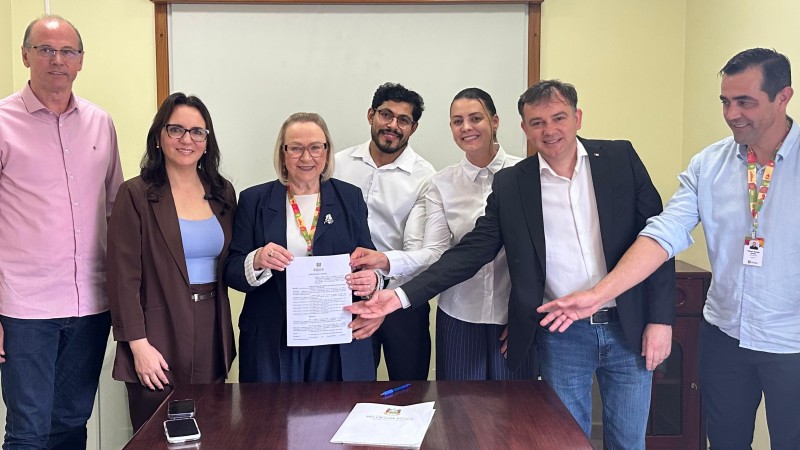Sete pessoas est&atilde;o posicionadas atr&aacute;s de uma mesa de madeira em uma sala de reuni&otilde;es. No centro do grupo, uma pessoa segura um documento impresso enquanto outras pessoas ao redor apontam ou apoiam as m&atilde;os pr&oacute;ximas ao documento, destacando o ato de assinatura ou formaliza&ccedil;&atilde;o. Sobre a mesa h&aacute; uma caneta, uma pasta com documentos e dois telefones celulares. Ao fundo, h&aacute; uma parede clara com um quadro branco emoldurado em madeira. As pessoas vestem roupas sociais ou esporte fino, em tons neutros e claros. O ambiente &eacute; iluminado por luz artificial interna.