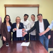 Sete pessoas est&atilde;o posicionadas atr&aacute;s de uma mesa de madeira em uma sala de reuni&otilde;es. No centro do grupo, uma pessoa segura um documento impresso enquanto outras pessoas ao redor apontam ou apoiam as m&atilde;os pr&oacute;ximas ao documento, destacando o ato de assinatura ou formaliza&ccedil;&atilde;o. Sobre a mesa h&aacute; uma caneta, uma pasta com documentos e dois telefones celulares. Ao fundo, h&aacute; uma parede clara com um quadro branco emoldurado em madeira. As pessoas vestem roupas sociais ou esporte fino, em tons neutros e claros. O ambiente &eacute; iluminado por luz artificial interna.