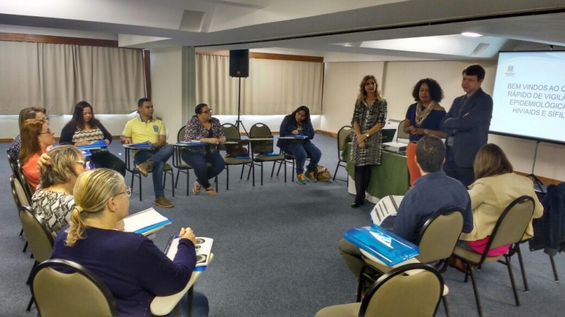 Três palestrantes (duas mulheres e um homem) estão de pé, dando as suas explicações enquanto a plateia está sentada, ouvindo atentamente.