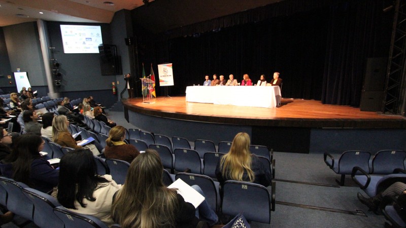 Sete participantes do evento estão sentados atrás de uma bancada no palco. O público, sentado, ouve atento. 