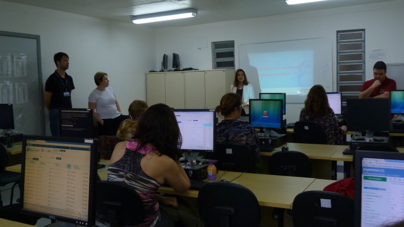 Palestrante, em pé, ao fundo da sala, dá explicações. Técnicos, sentados, trabalham em seus computadores.