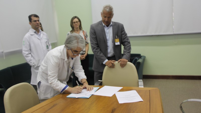 Nadine, abaixada, assina documento observada pelo secretário Gabbardo, que está apoiado numa cadeira. Atrás deles, um profissional de saúde e uma mulher. 