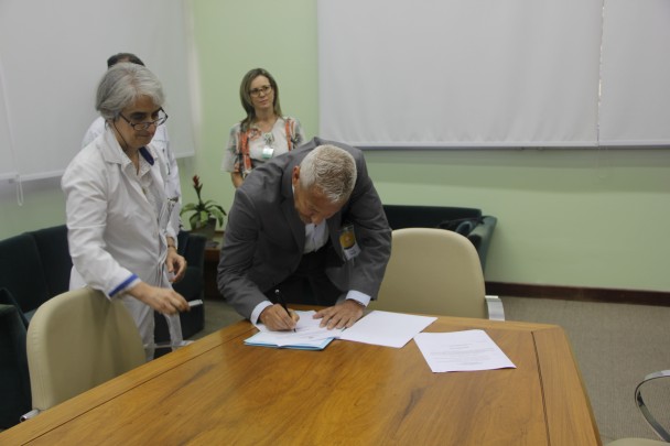 Secretário Gabbardo, abaixado, assina documento apoiado numa mesa. Atrás deles, um profissional de saúde e uma mulher.