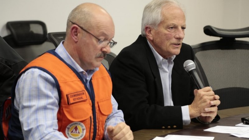 Vice-governador Cairoli fala com o microfone na mão. Um homem está ao seu lado. Os dois estão sentados atrás de uma mesa.