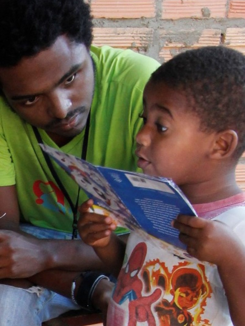 Criança atendida no Programa Primeira Infância Melhor de Viamão olha com atenção um livro infantil sob a supervisão atenta do visitador integrante do programa