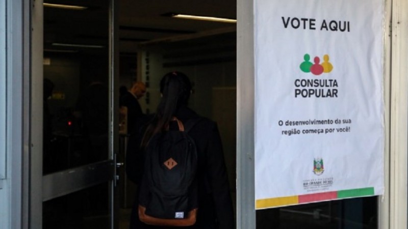 Porta aberta com banner escrito "Consulta Popular". No fundo, uma pessoa de mochila.