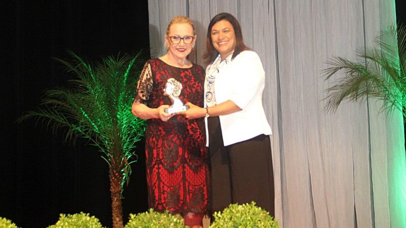 Secretária Arita, de pé, está ao lado da mestre de cerimônias. As duas seguram o troféu recebido pela secretária.