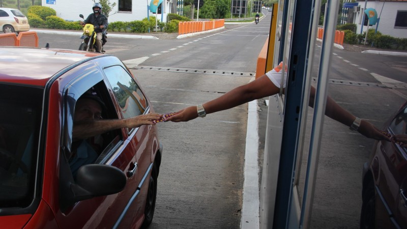 Motorista de carro vermelho estica o braço para fora da janela do veículo para receber preservativo que está sendo entregue por uma funcionária do pedágio.
