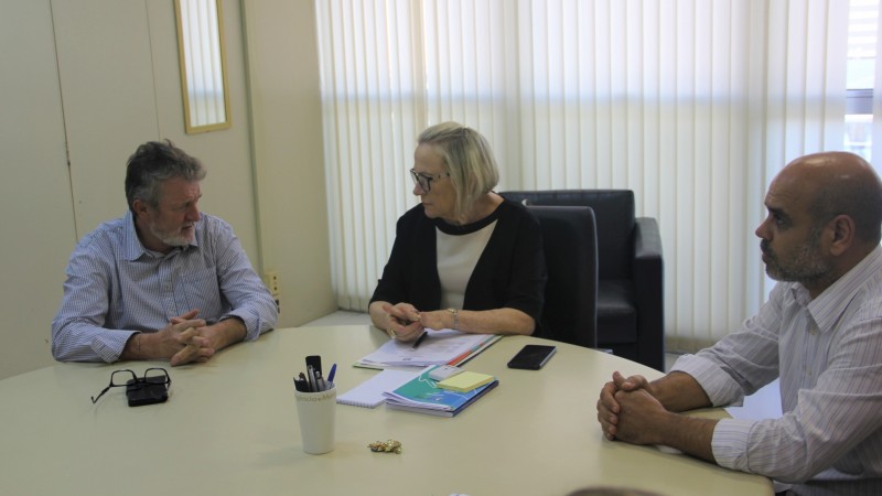 Secretária Arita (no centro) ouve a explicação do participante da reunião que está a sua esquerda. O outro participante está a sua direita. Todos estão sentados. 