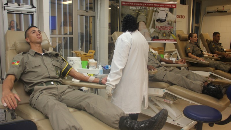 Quatro pessoas com farda da Brigada Militar, sentadas em cadeiras de doação de sangue e com o braço esquerdo estendido. Também aparece uma enfermeira, de jaleco branco, virada de costas.
