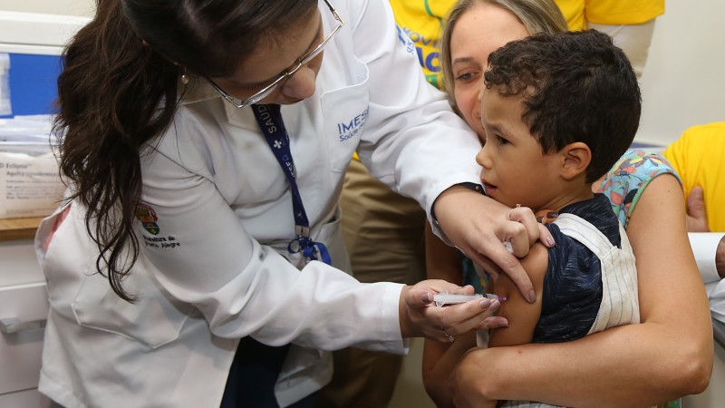 A imagem mostra um menino sendo vacinado por uma profissional. Uma outra mulher o segura enquanto ele faz cara de choro. 