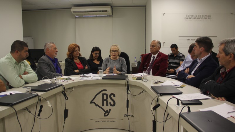 Secretária Arita fala enquanto participantes da reunião ouvem atentamente. Todos estão sentados em volta de uma mesa.