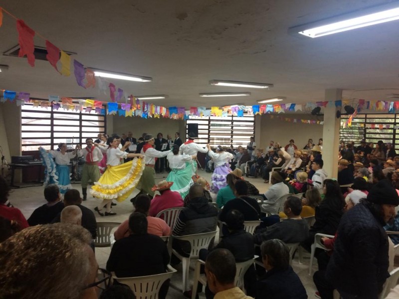 Grupo de dança tradicionalista gaúcho se apresenta, com bandeirinhas penduradas no teto e plateia sentada assistindo.
