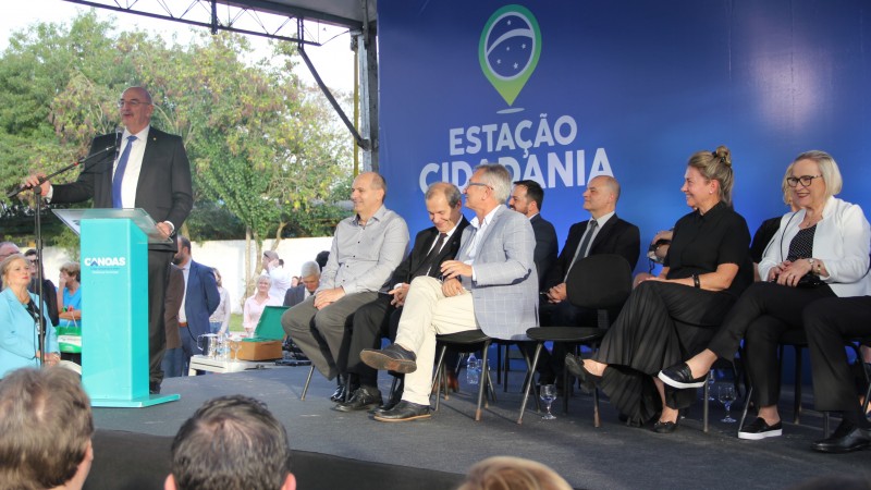 Ministro discursa de pé, atrás de um púlpito, enquanto algumas autoridades assistem sentadas. A plateia está a frente.  