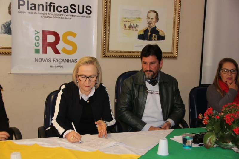 Secretária Arita, sentada, assinando um documento enquanto um homem e uma mulher a observam.