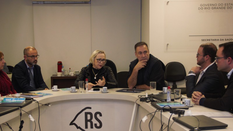 Arita fala para participantes da reunião. Todos, homens e mulheres, estão sentados em volta de uma mesa.