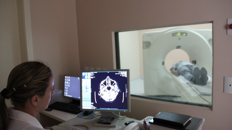 Uma médica observa, através de uma tela de computador e de um vidro, uma pessoa realizando uma tomografia.