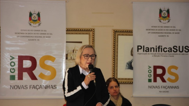 Secretária Arita de pé, falando ao microfone. À esquerda da foto, atrás da secretária, há um banner escrito "GOV RS novas façanhas", e à direita da foto, outro banner escrito "PlanificaSUS".