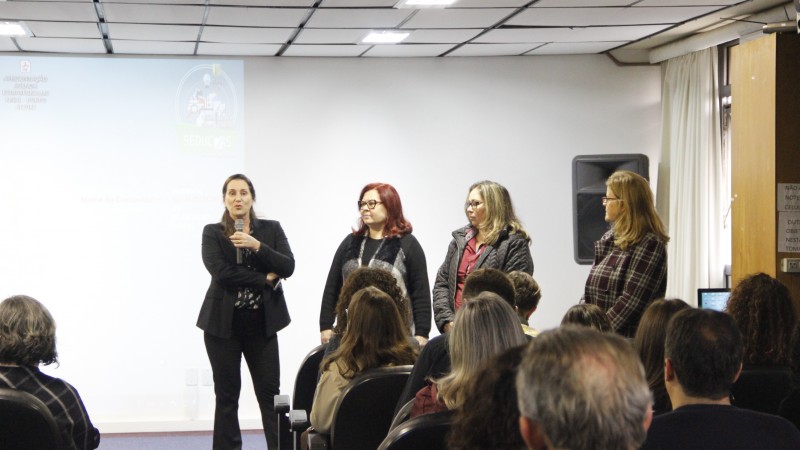As quatro palestrantes estão de pé. Uma fala com o microfone na mão. A plateia assiste sentada. 