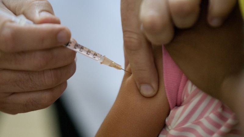 A imagem mostra a mão de um médico dando vacinando o braço de uma menina.